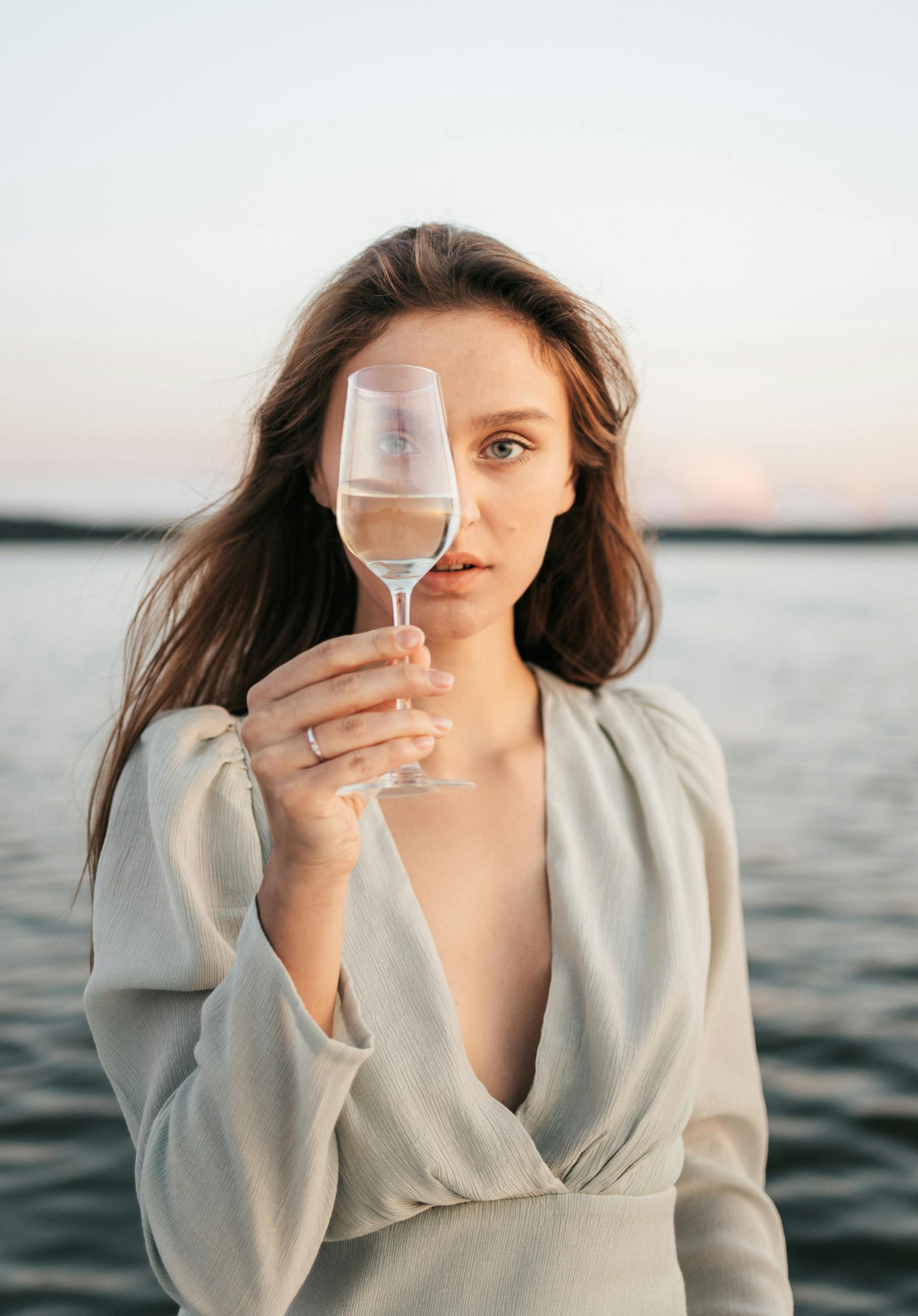 Elegant woman holding a champagne glass by a serene lake at twilight, creating a peaceful and sophisticated atmosphere.