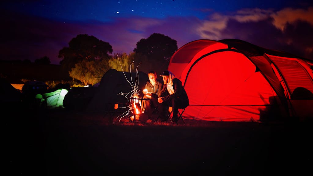 Two men enjoy a campfire by a red tent under a starry sky in Scotland.